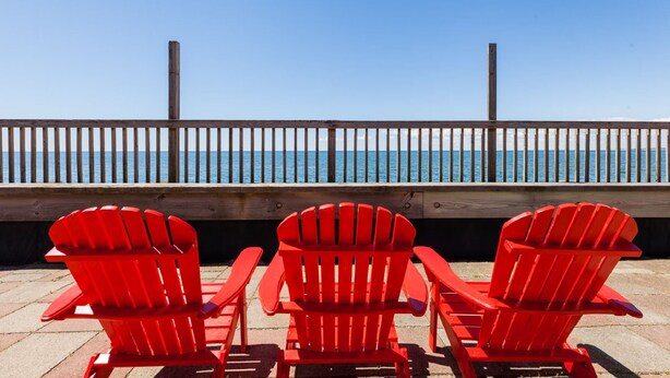 Tres sillas Adirondack rojas mirando a una terraza de madera con barandilla y vista a la playa.