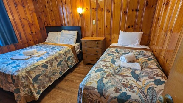 Une chambre avec deux lits superposés en bois, chacun recouvert d’une courtepointe à motifs floraux et agrémenté d’oreillers blancs. Une commode en bois est placée entre les lits, et un luminaire mural est fixé au-dessus.