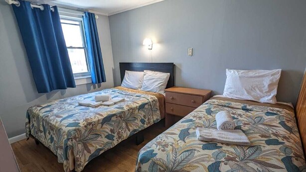 Una habitación con dos camas, una mesita de noche, una ventana con cortinas y una lámpara en la pared.