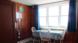 A dining area with a wooden table and chairs, a window with curtains, and a door with a fire extinguisher. - Unwind on the Oceanside in the BlueFish Saturday Cottage at Oceana Cottages (North Truro)