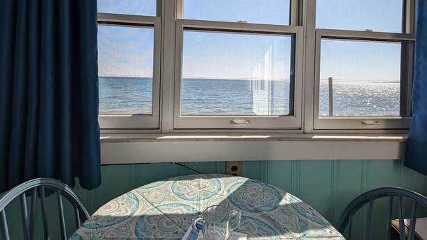 A room with a table covered by a patterned tablecloth, three windows with a view of the sea, and a chair.
