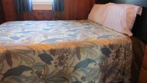 A bed with a floral bedspread and a single white pillow in a room with wooden walls and a window with curtains.