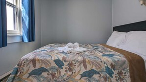 A bedroom with a bed, a window with curtains, a wall-mounted light fixture, and a headboard. - Tranquil Getaway at the Dolphin Sunday cottage located at the Oceana Cottages (North Truro)