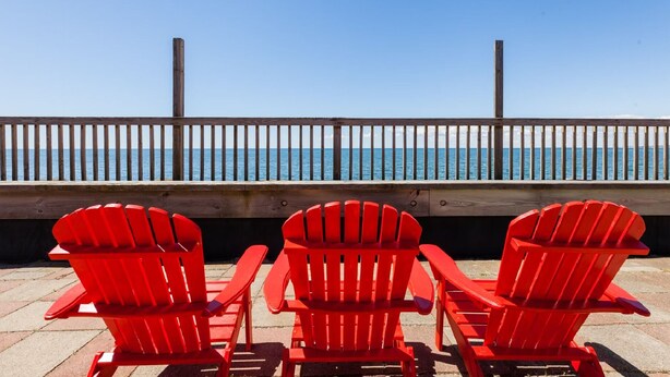 Three red Adirondack chairs facing a wooden deck with a railing overlooking a beach. - Tranquil Getaway at the Dolphin Sunday cottage located at the Oceana Cottages (North Truro)
