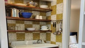 A compact kitchen with wooden cabinets, a sink, and shelves above holding dishes and utensils. - Relaxing Retreat at the Dolphin Saturday cottage located at the Oceana Cottages (North Truro)