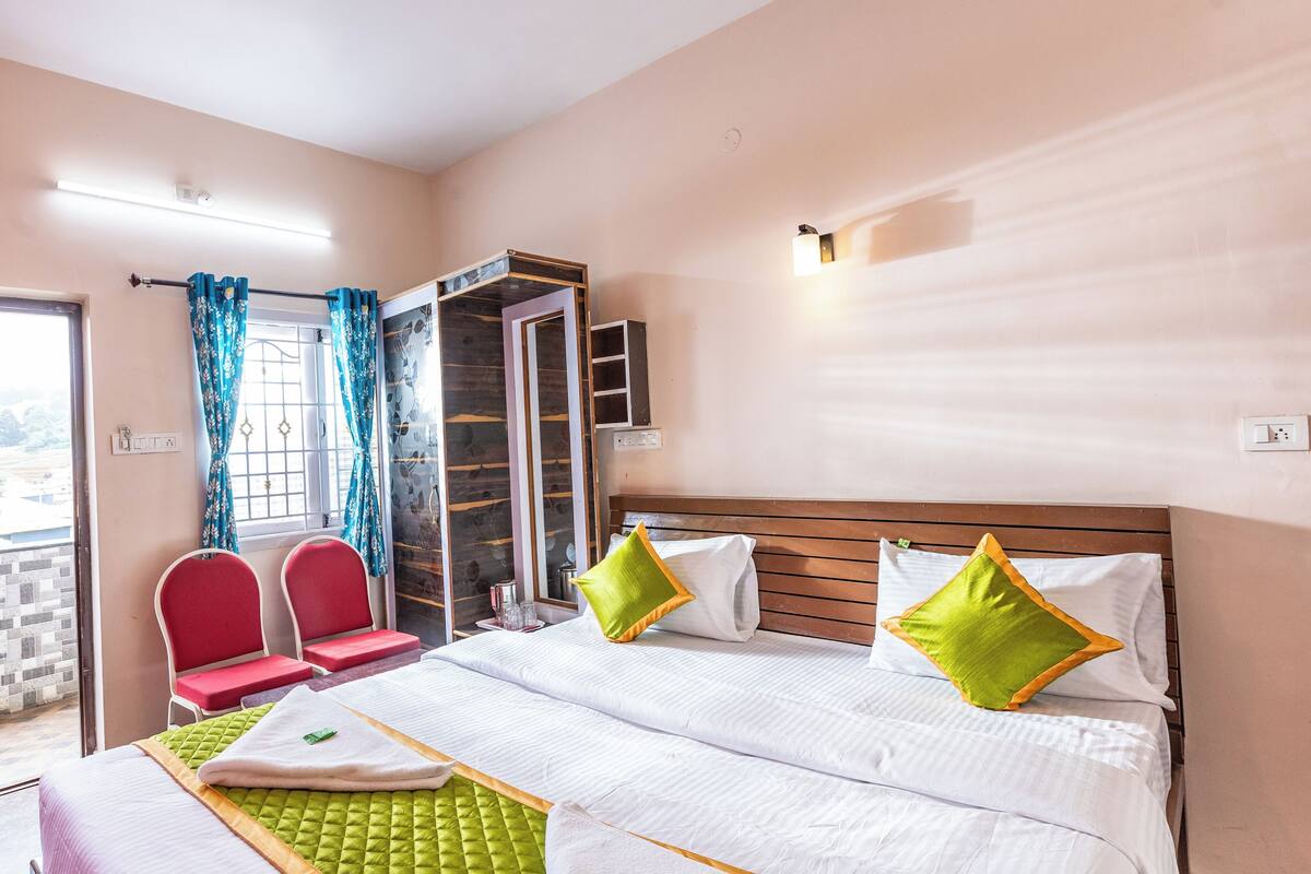 A hotel room with a bed, two red chairs, a small table, a mirror, and a window with blue curtains.