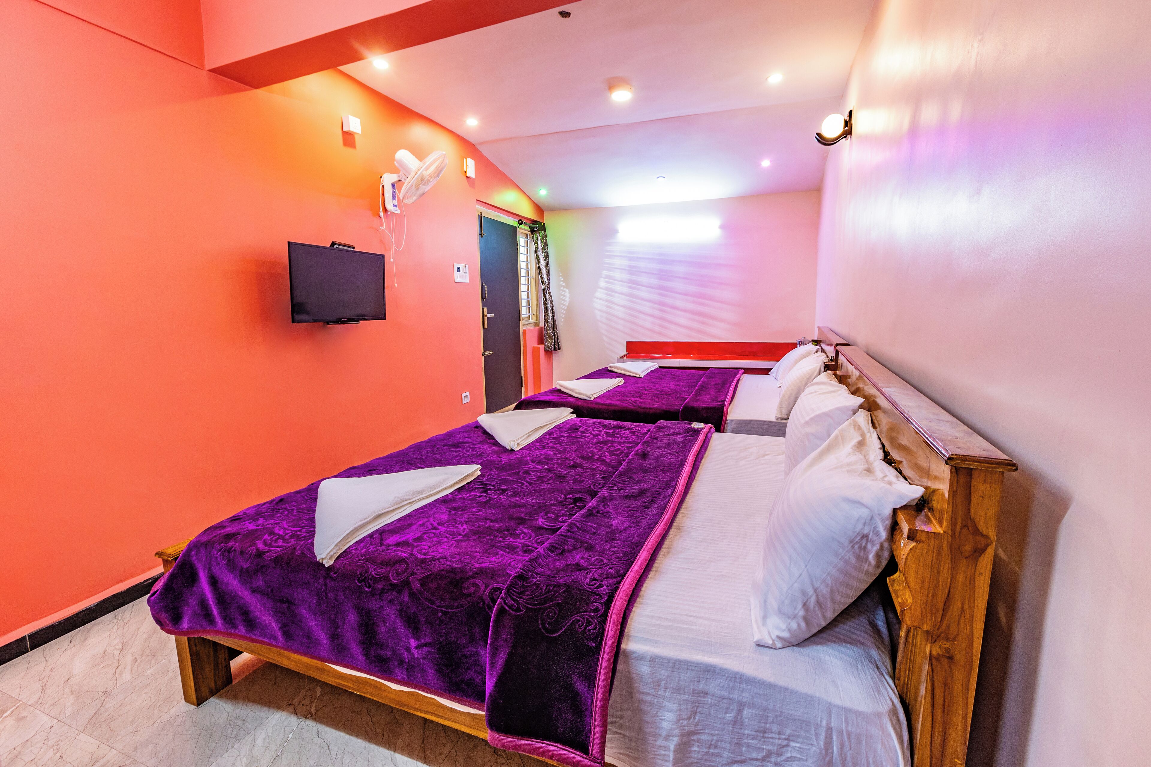 A hotel room with two beds, each with a purple blanket and white pillows, a flat-screen TV mounted on the wall, and a ceiling fan.