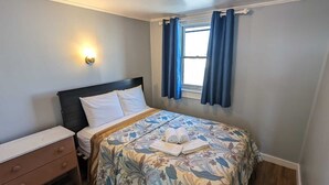 Un dormitorio con cama, mesita de noche, cómoda, ventana con cortinas y lámpara de pared.
