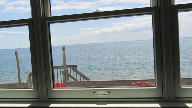 Une vue à travers une fenêtre donnant sur une terrasse en bois et une balustrade, avec une mer calme en contrebas.