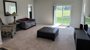 A spacious living room with a grey sofa, a black ottoman, a lamp, and a large window offering a view of a grassy yard.