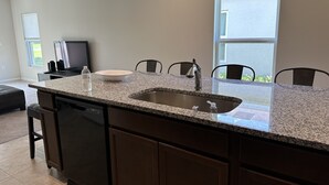 A kitchen with granite countertops, a stainless steel sink, and a dishwasher. There is a dining area with chairs and a table.