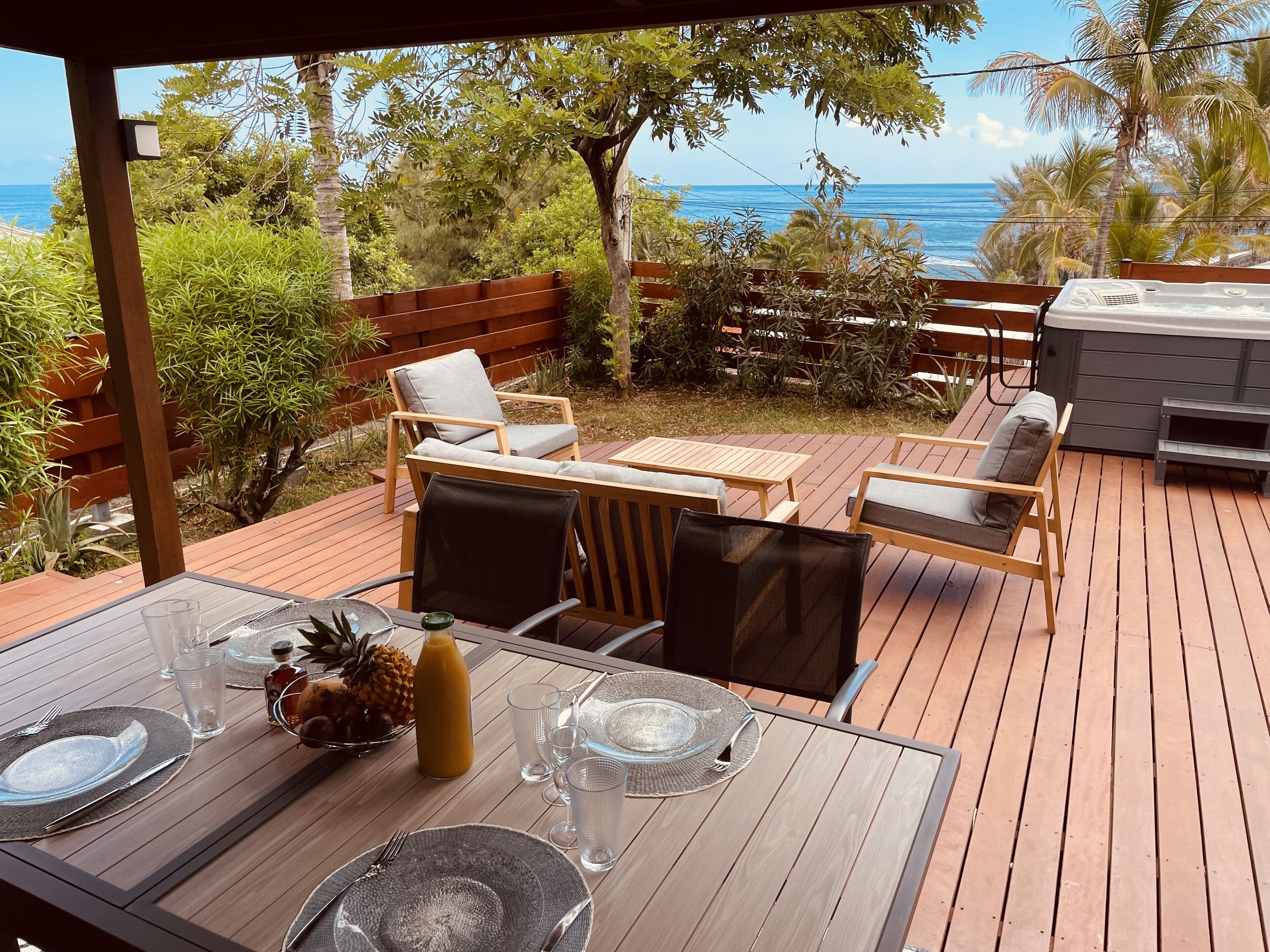 Une terrasse en bois aménagée avec du mobilier d’extérieur, notamment une table et des chaises pour les repas, donnant sur une plage bordée de palmiers et sur l’océan.