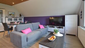 A modern living room with a grey sofa, a dining table with chairs, a flat-screen TV, and a kitchen area with cabinets and a window. - Comfortable apartment in Burg-Reuland with shared terrace (Burg-Reuland)