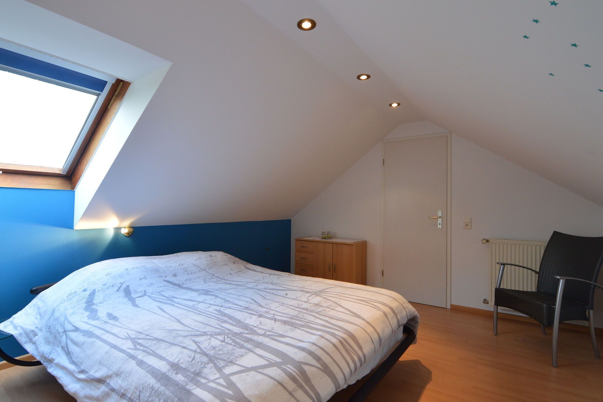 Une chambre avec un plafond en pente, un puits de lumière, un lit recouvert d’une couvre-lit à motifs, une armoire en bois, une chaise et un radiateur.