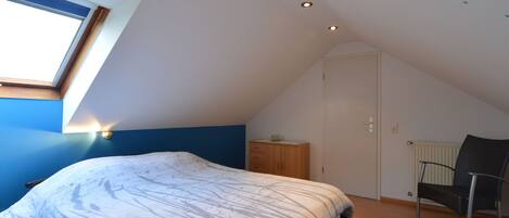Une chambre avec un plafond en pente, un puits de lumière, un lit recouvert d’une couvre-lit à motifs, une armoire en bois, une chaise et un radiateur.