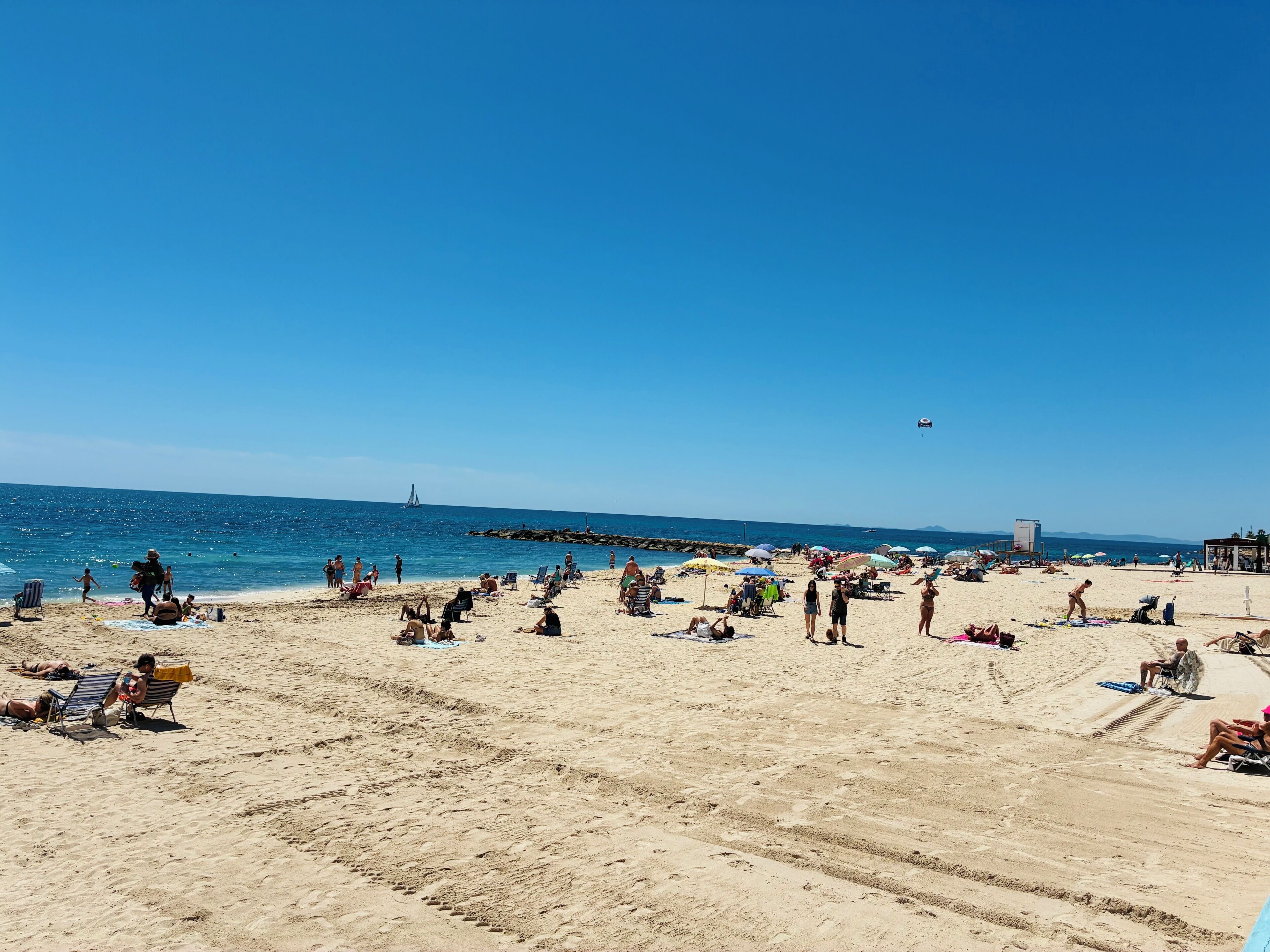 En strand med folk, der solbader og svømmer, en sejlbåd i det fjerne, og et redderhus.