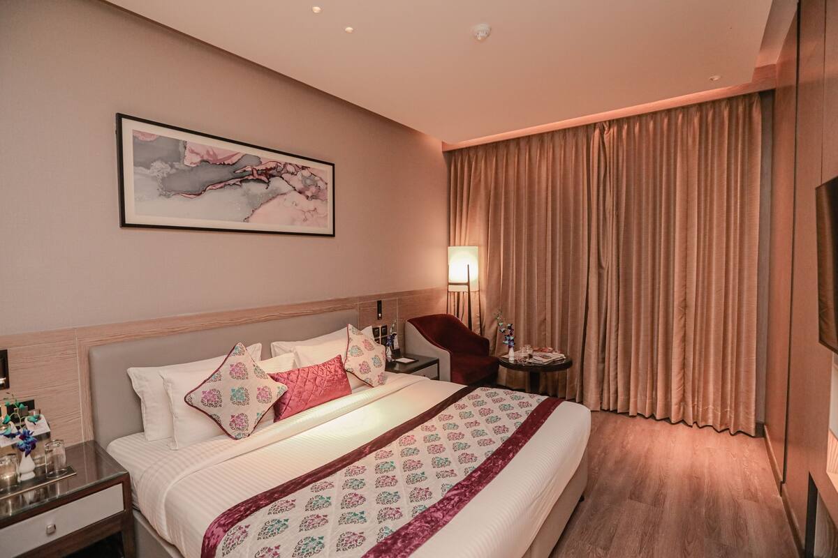 A hotel room with a large bed, a nightstand, a chair, a lamp, and a painting on the wall.