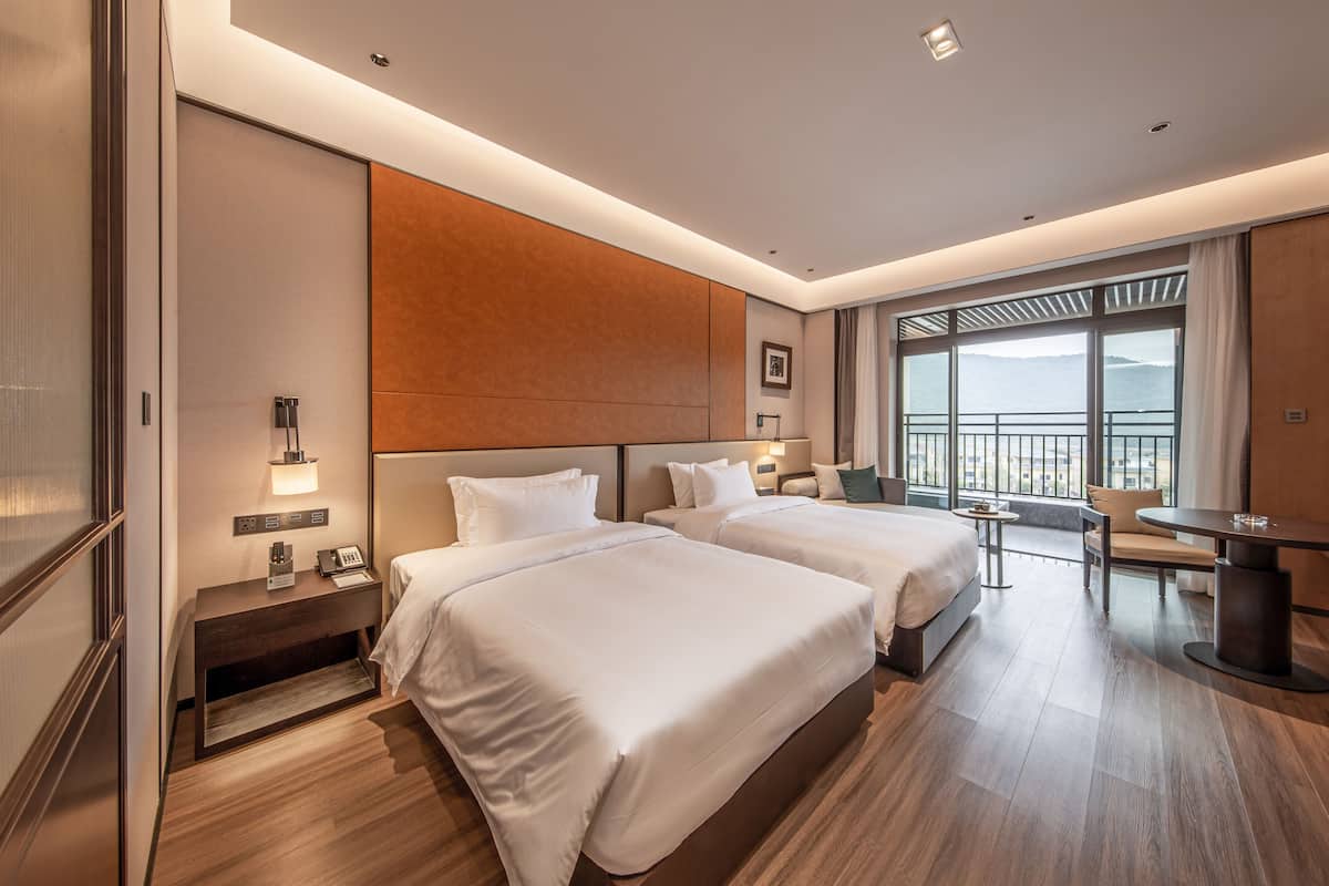 A hotel room with two beds, a desk, a chair, a small table, a nightstand, a lamp, and a large window with a view.