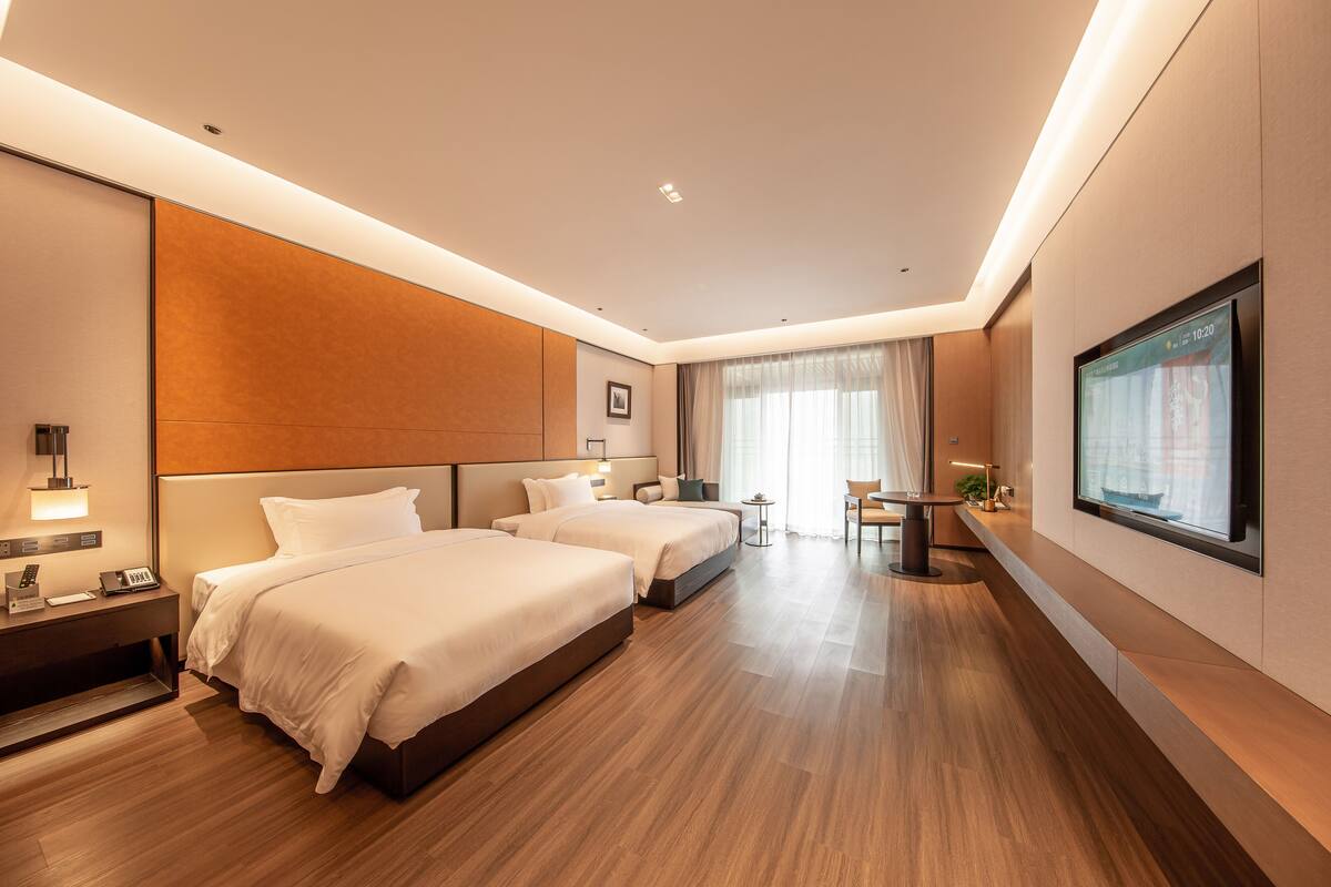 A modern hotel room with two beds, a desk, a chair, and a TV on the wall.