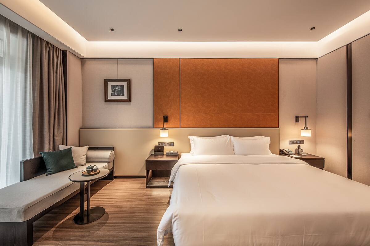 A modern hotel room with a large bed, a sofa, a small table, and two bedside tables.