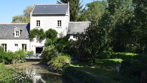 Un bâtiment blanc surmonté d’un toit gris, entouré d’une végétation luxuriante et bordé d’une étendue d’eau paisible.
