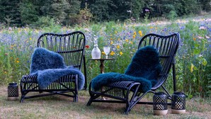 Deux fauteuils en osier noir, recouverts de plaids en fausse fourrure bleue, une petite table avec une bouteille et un verre, et, en arrière-plan, un champ de fleurs sauvages.