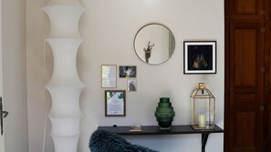 A room with a white wall, a round mirror, framed pictures, a small table with a green vase and a candle, and a dark blue furry object.