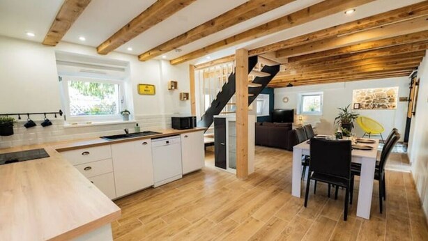 A modern kitchen with wooden beams, a dining area with a black chair, and a staircase leading to another level. - Charming renovated gîte (Rimbach-prés-Guebwiller)