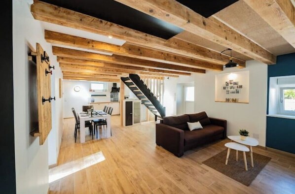 Amplia sala de estar con una escalera que conduce a una cocina, vigas de madera en el techo y un comedor con mesa y sillas.