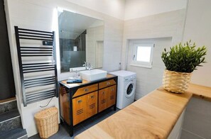 Un baño moderno con tocador de madera, lavadora integrada, un espejo grande y un ventanal pequeño.