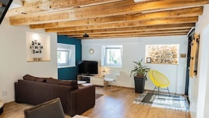 A cozy living room with a brown sofa, a dining area with a table and chairs, a TV, and a stone wall. - Charming renovated gîte (Rimbach-prés-Guebwiller)