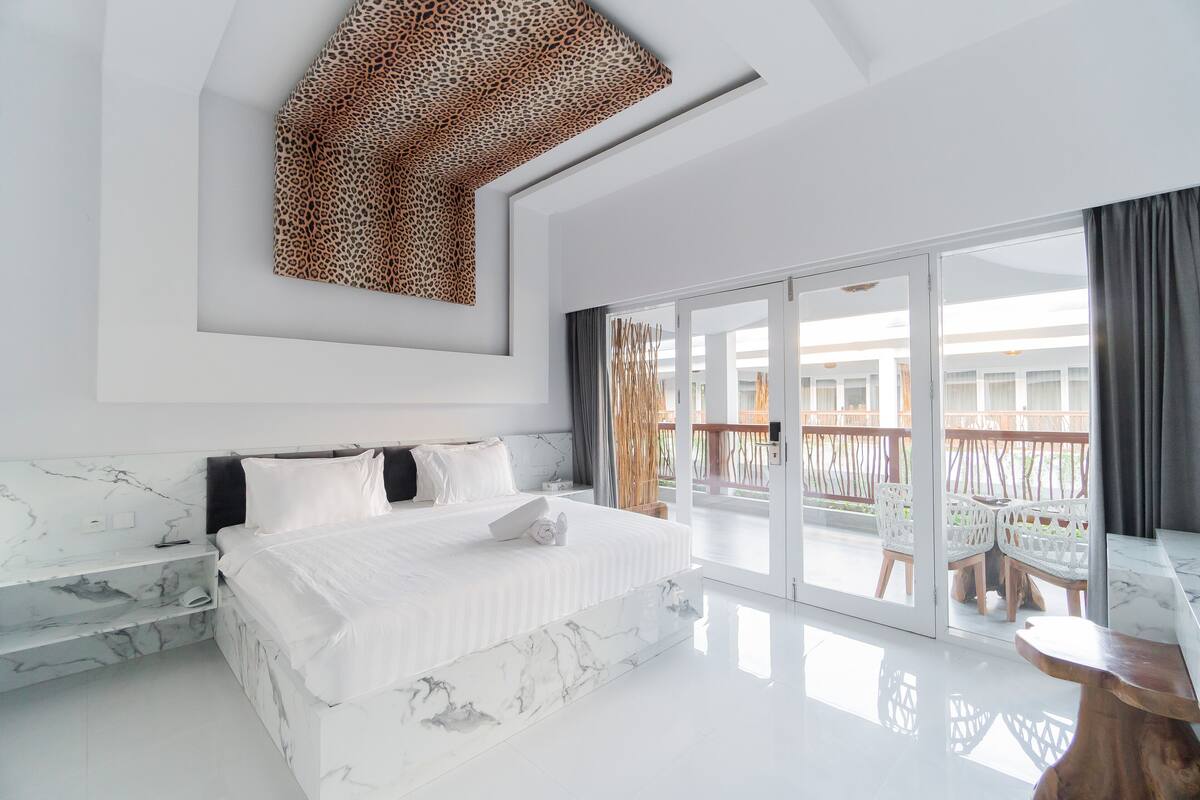 Une chambre moderne dotée d’un grand lit, d’éléments décoratifs en marbre, d’une œuvre d’art au plafond et d’un balcon aménagé avec du mobilier d’extérieur.
