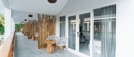 A covered outdoor seating area with a long table and chairs, a hanging light fixture, and a view through large glass doors.