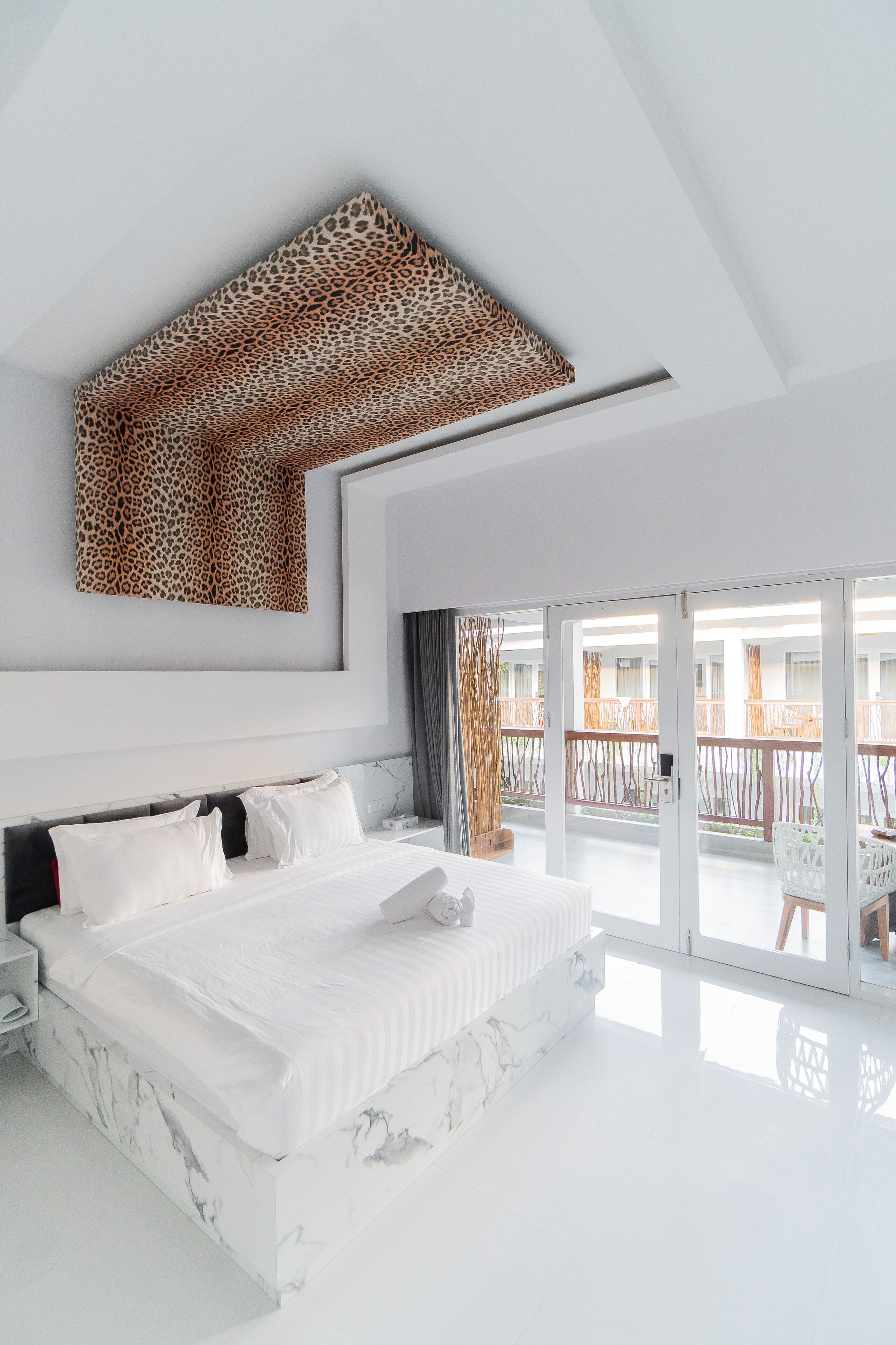 A modern bedroom with a large bed, a ceiling artwork, and a balcony with outdoor furniture.