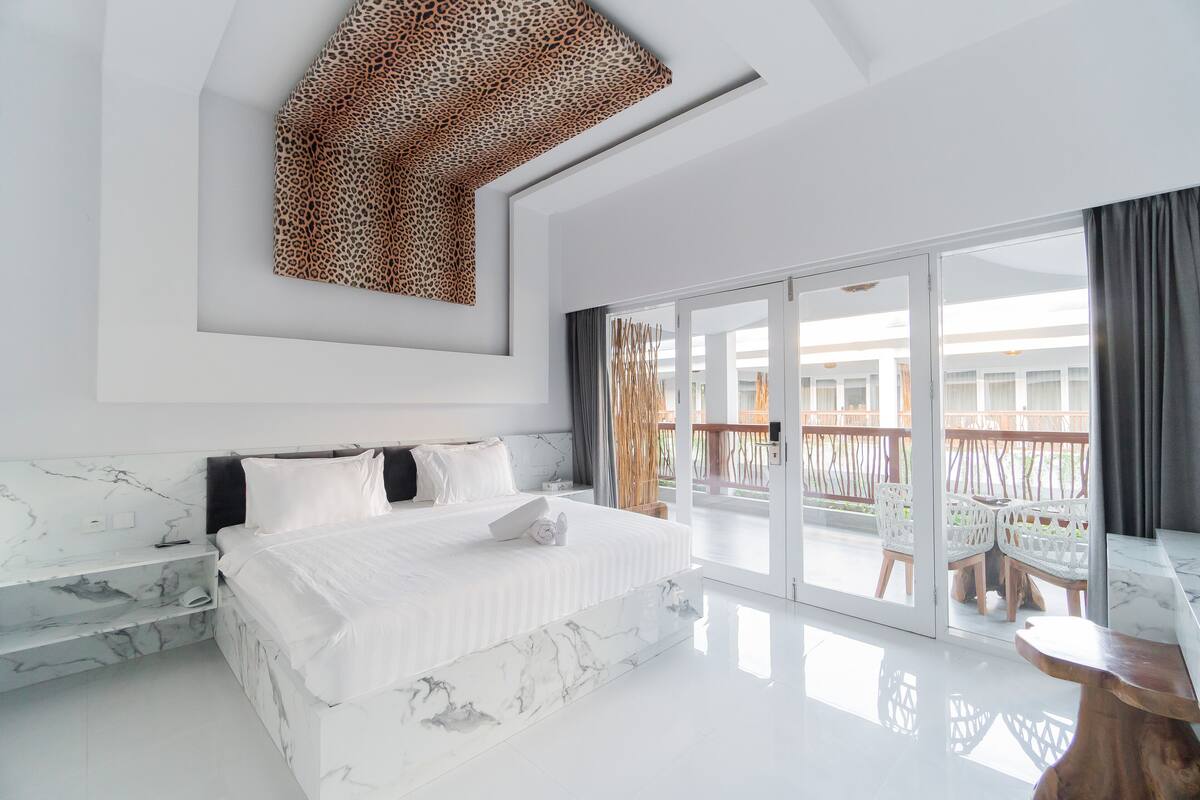 Une chambre d’hôtel moderne dotée d’un grand lit, d’éléments décoratifs en marbre, d’un balcon avec des meubles en osier et d’une œuvre d’art murale à motifs léopard.