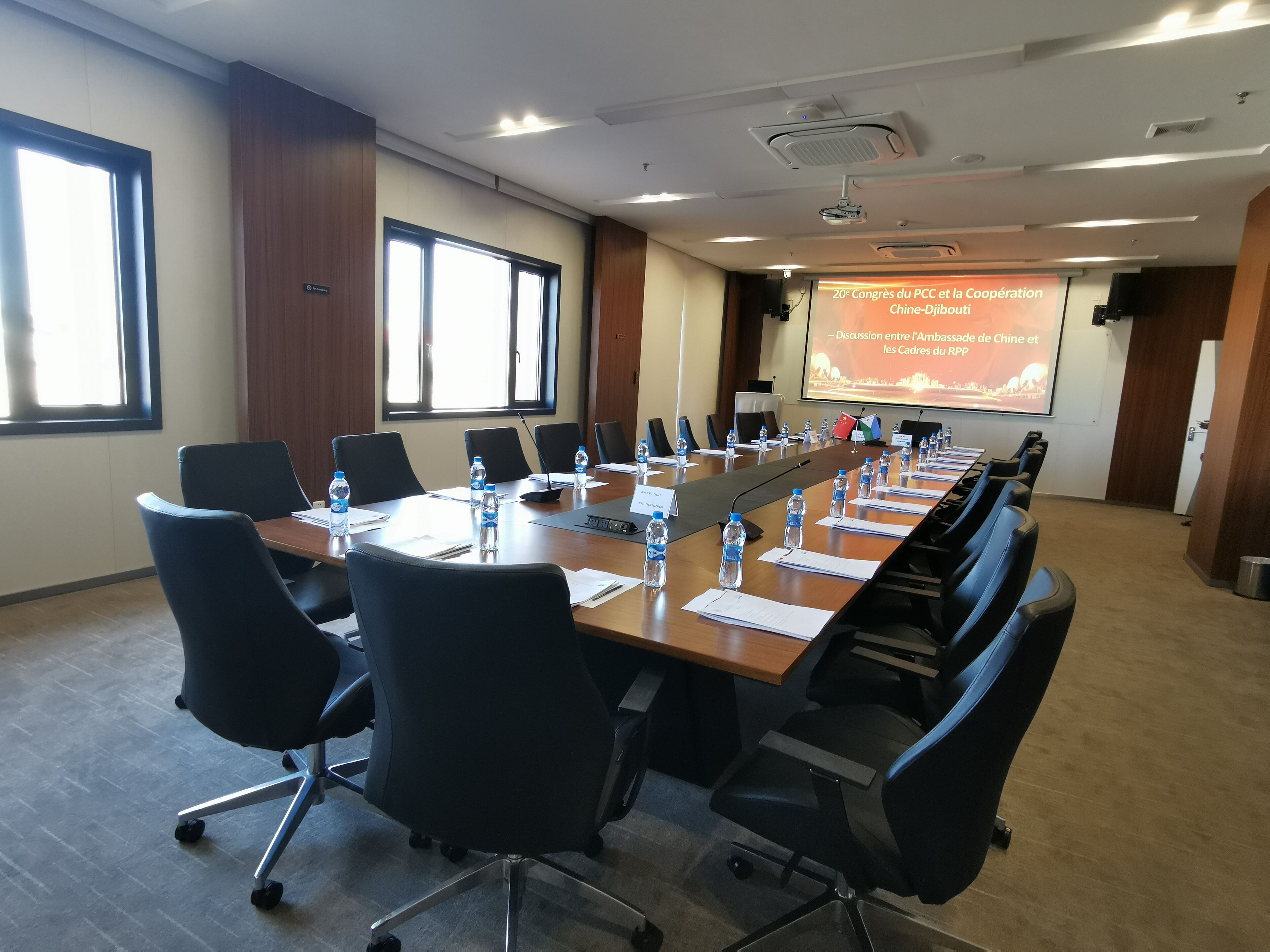 A conference room with a long table, black chairs, water bottles, and a projector screen displaying a presentation.