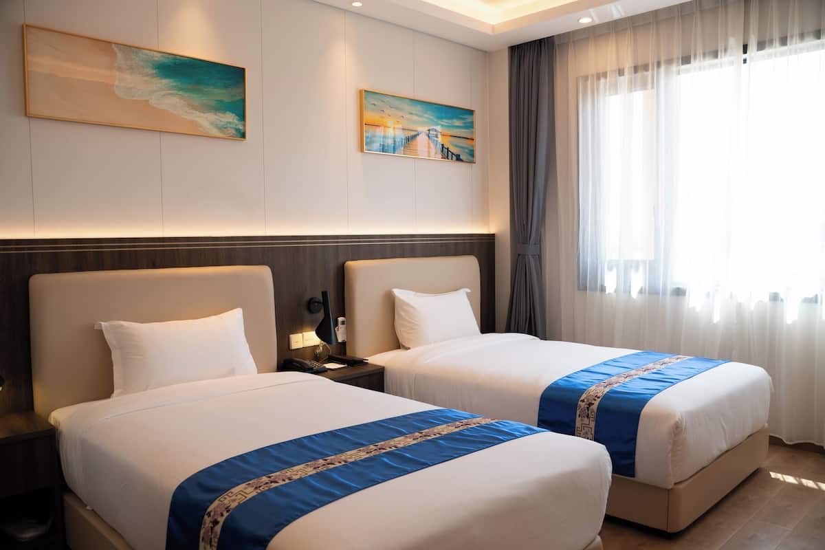 A hotel room with two beds, a nightstand, a painting, and a window with curtains.