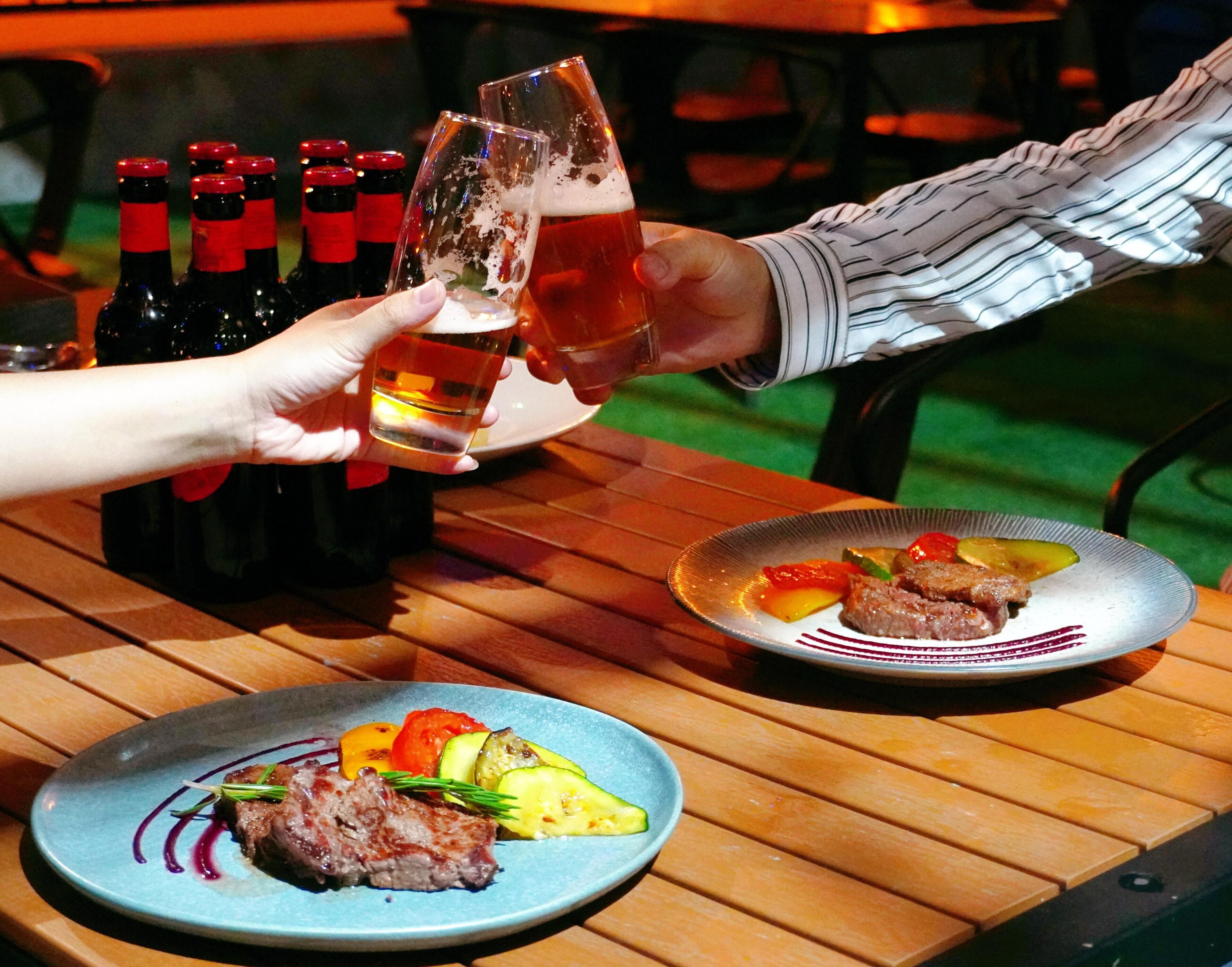 A dining table with two plates of food, a bottle of beer, and a person holding a glass.