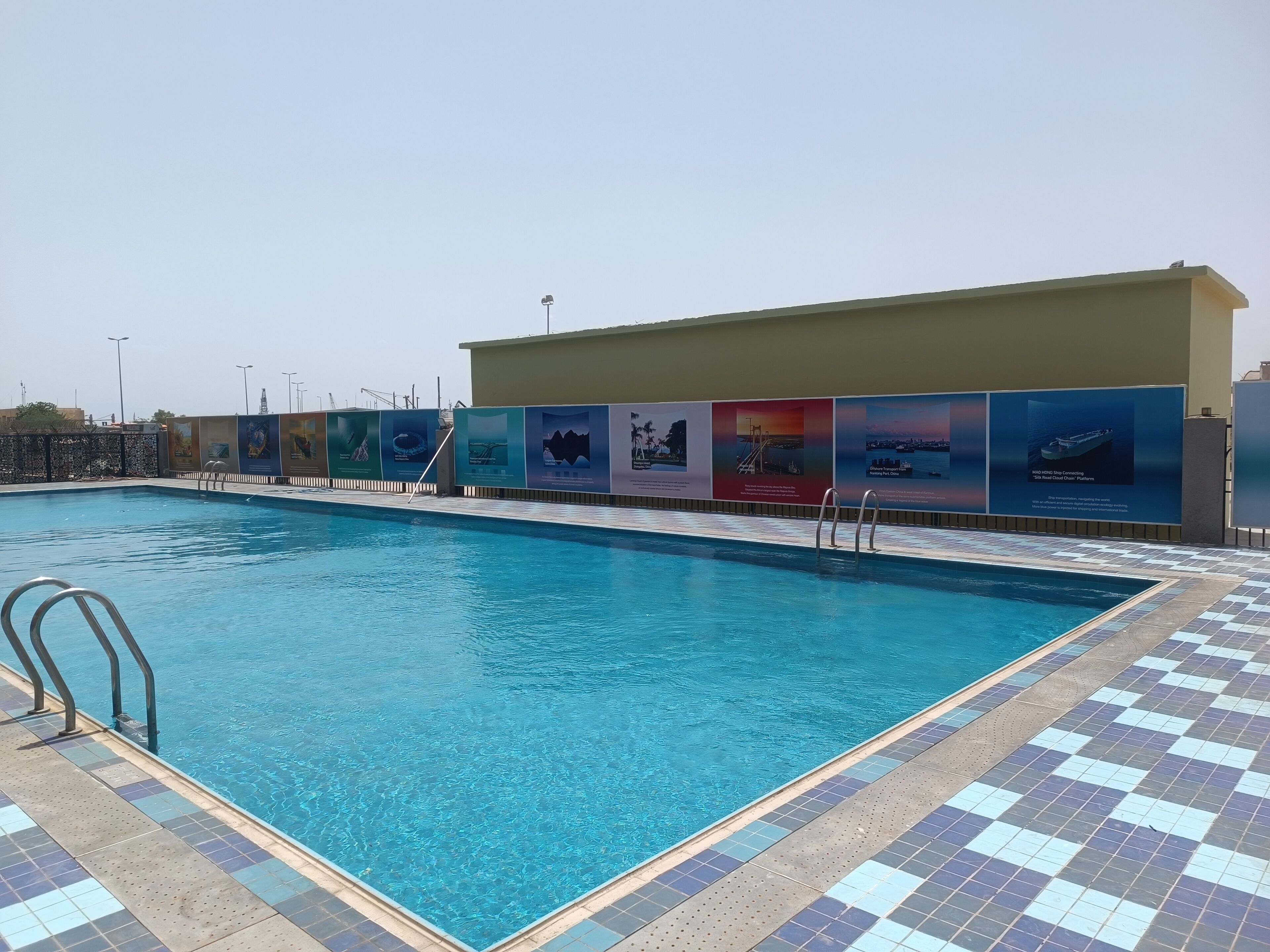 A swimming pool with tiled edges, a metal ladder, and a building with multiple advertisements.