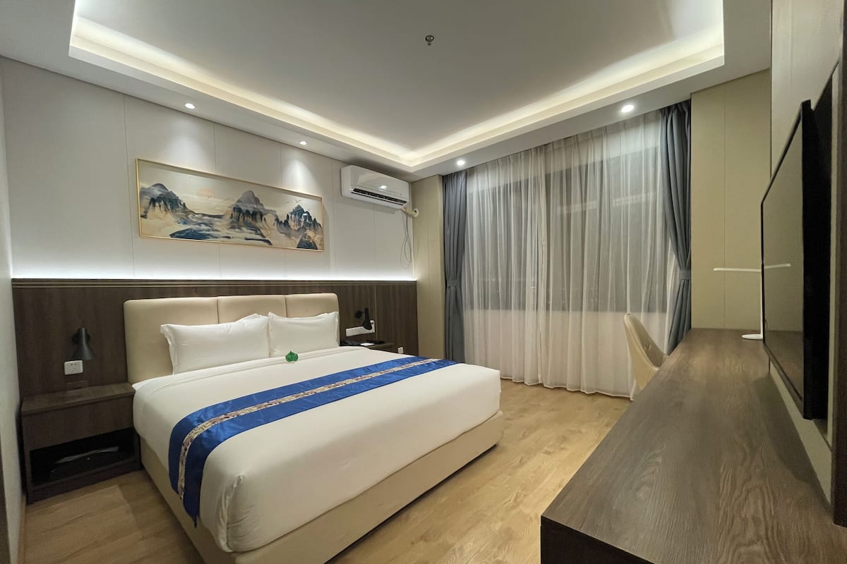 A modern hotel room with a large bed, a bedside table, a desk, a flat-screen TV, and a painting on the wall.