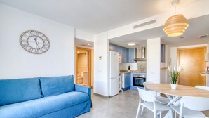 A modern kitchen with a built-in oven, a dining area with a round table and white chairs, a blue sofa, and a wall-mounted clock.