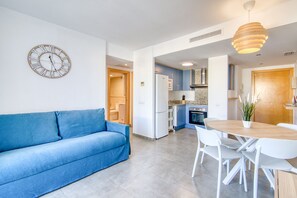 Une cuisine moderne dotée d’un four encastré, un coin repas avec une table ronde et des chaises blanches, un canapé bleu et une horloge murale.