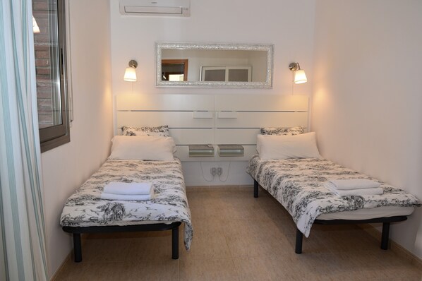 A room with two beds, each with a patterned bedspread and a white towel on the bed. There is a mirror above the beds and two wall-mounted lights.
