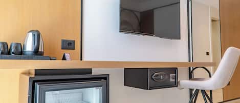 A compact room with a flat-screen TV mounted on the wall, a small refrigerator, a chair, and a coffee maker on the counter.