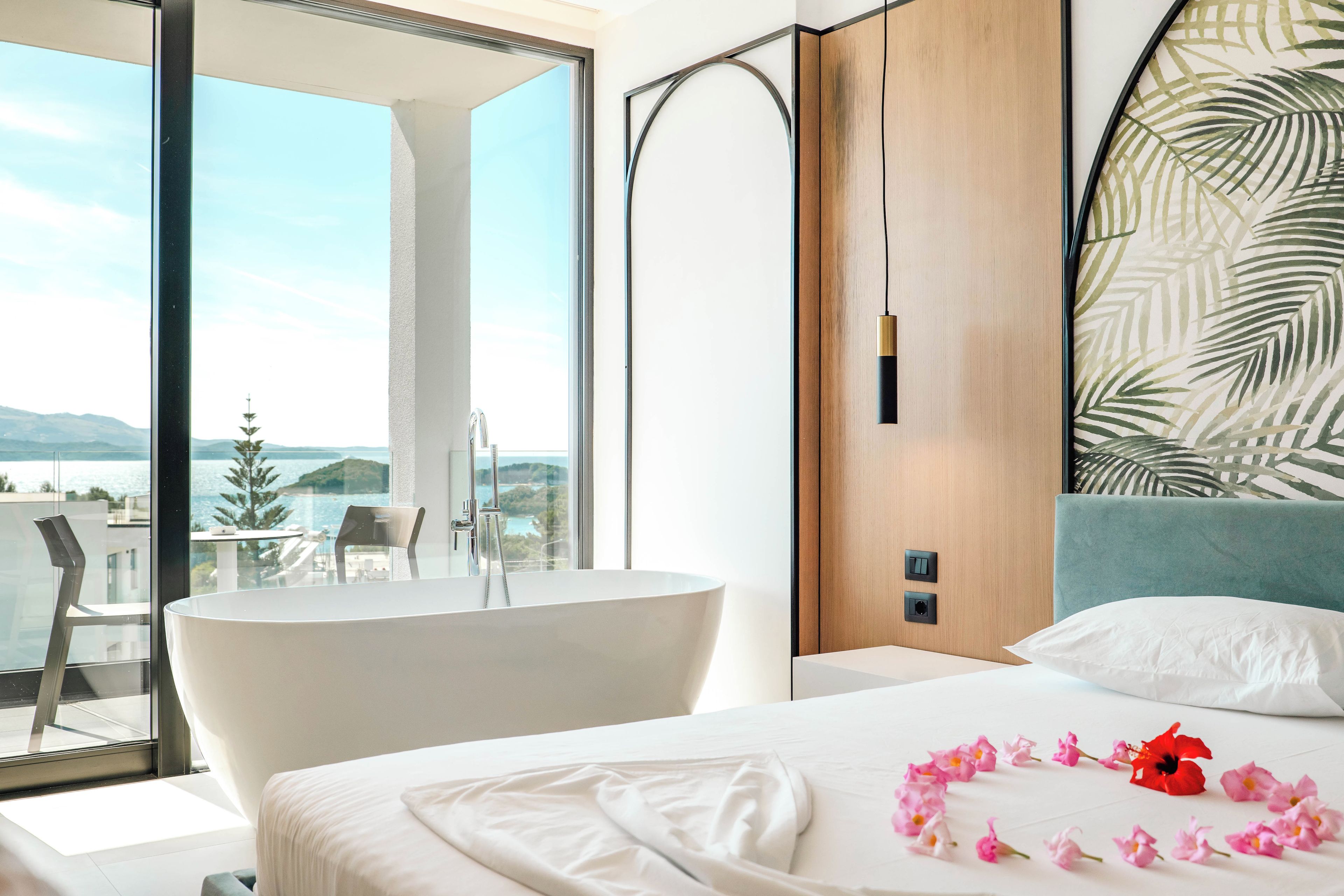 Ein modernes Hotelzimmer mit einem großen Fenster, einer Badewanne, einem Bett mit weißen Leinen und Blick auf das Meer.