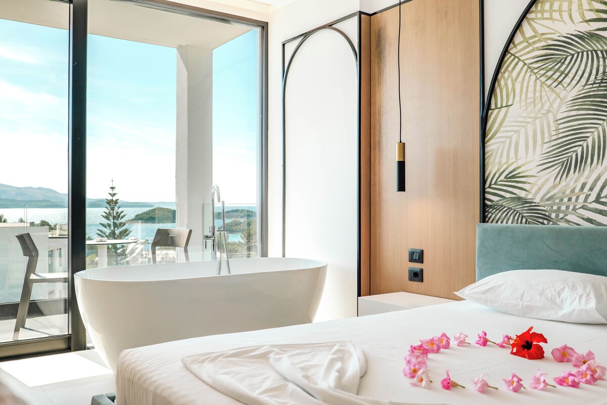 A modern hotel room with a large window, a bathtub, a bed with white linens, and a view of the sea.