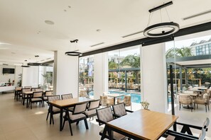 A modern indoor dining area with tables and chairs, large windows, and a view of an outdoor pool area. - Hotel Colosseum (Ksamil)
