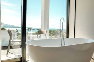 A modern bathroom with a freestanding bathtub, large windows, and a view of a body of water and mountains.