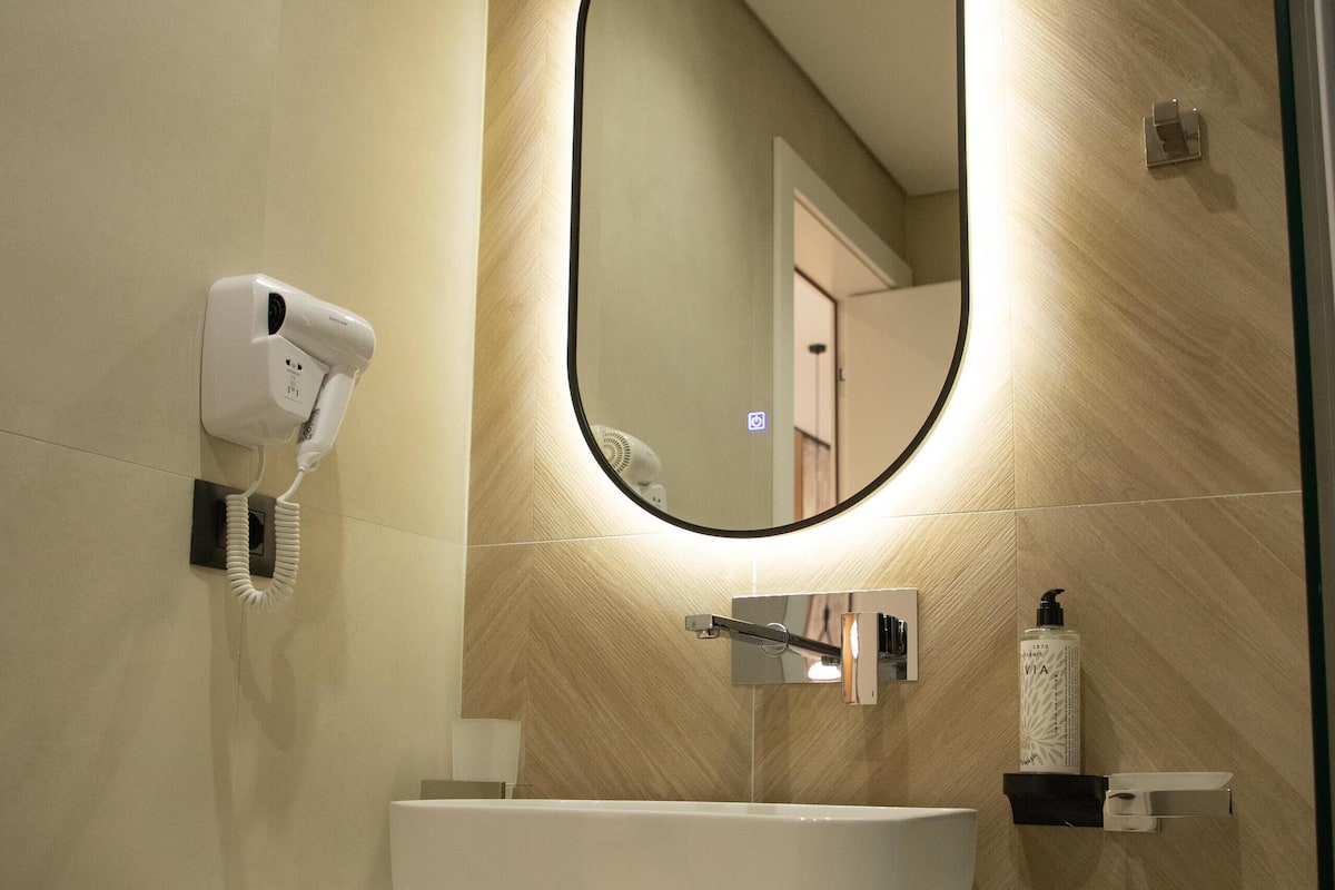 A modern bathroom with a white sink, a round mirror with a black frame, and a hairdryer mounted on the wall.