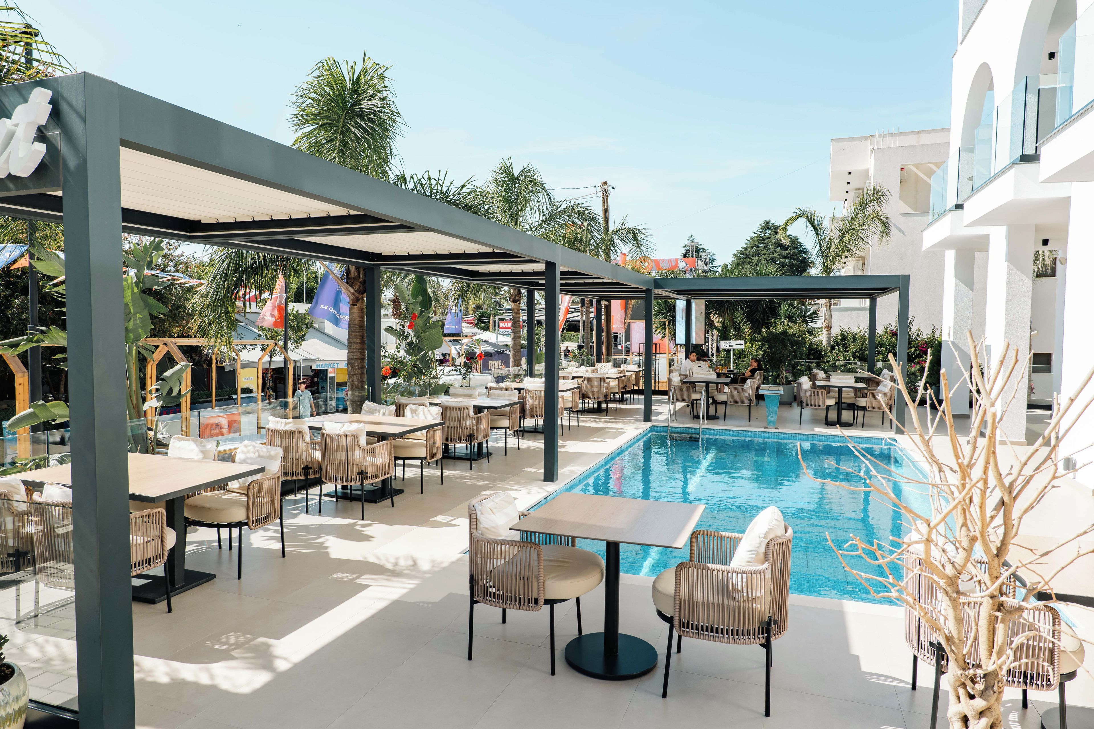 Área de piscina externa com espreguiçadeiras e mesas, rodeada por um espaço coberto com assentos e a própria piscina.